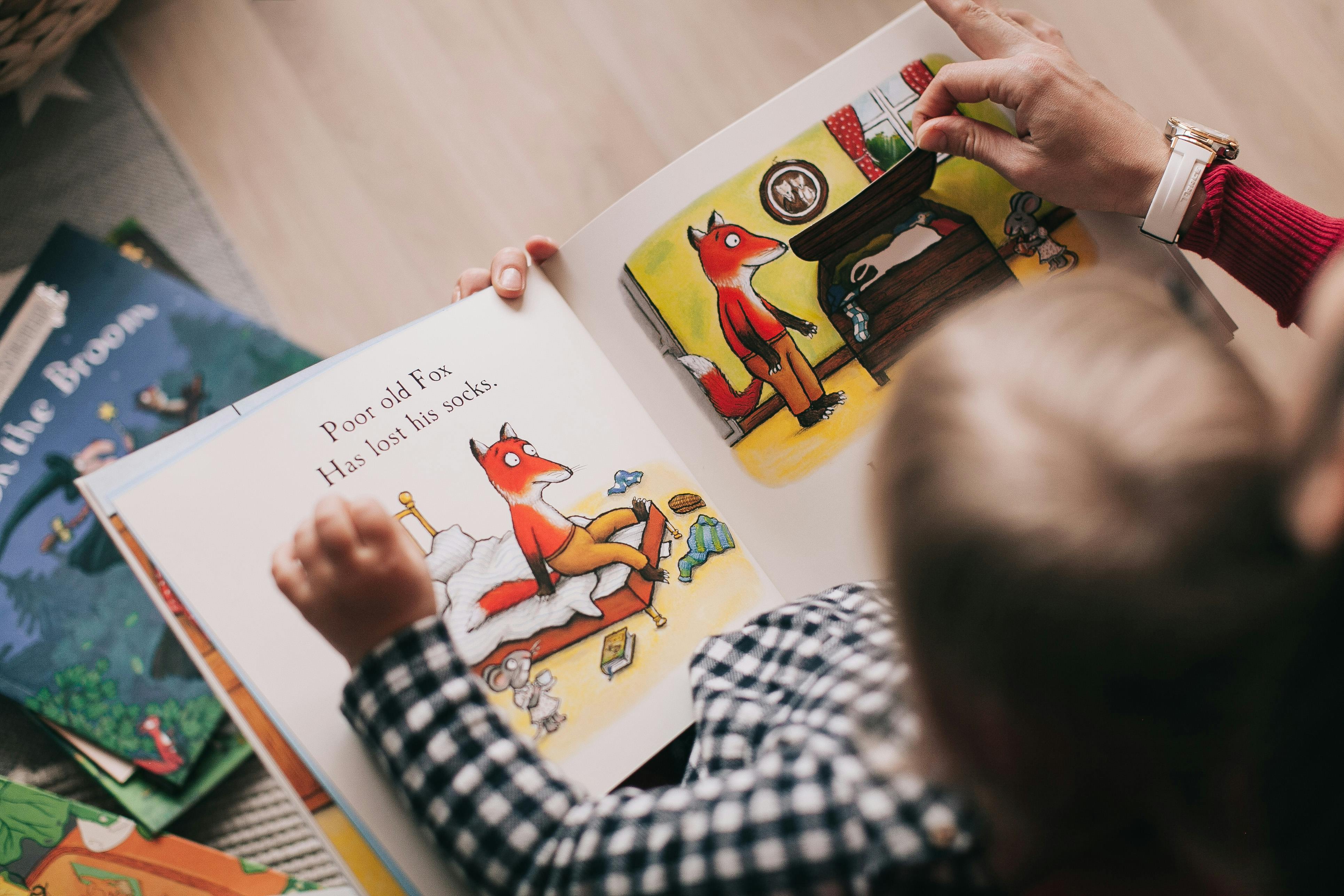 A person wearing a red sweater and a black and white plaid shirt is holding an open book with a picture of a fox lying in ...