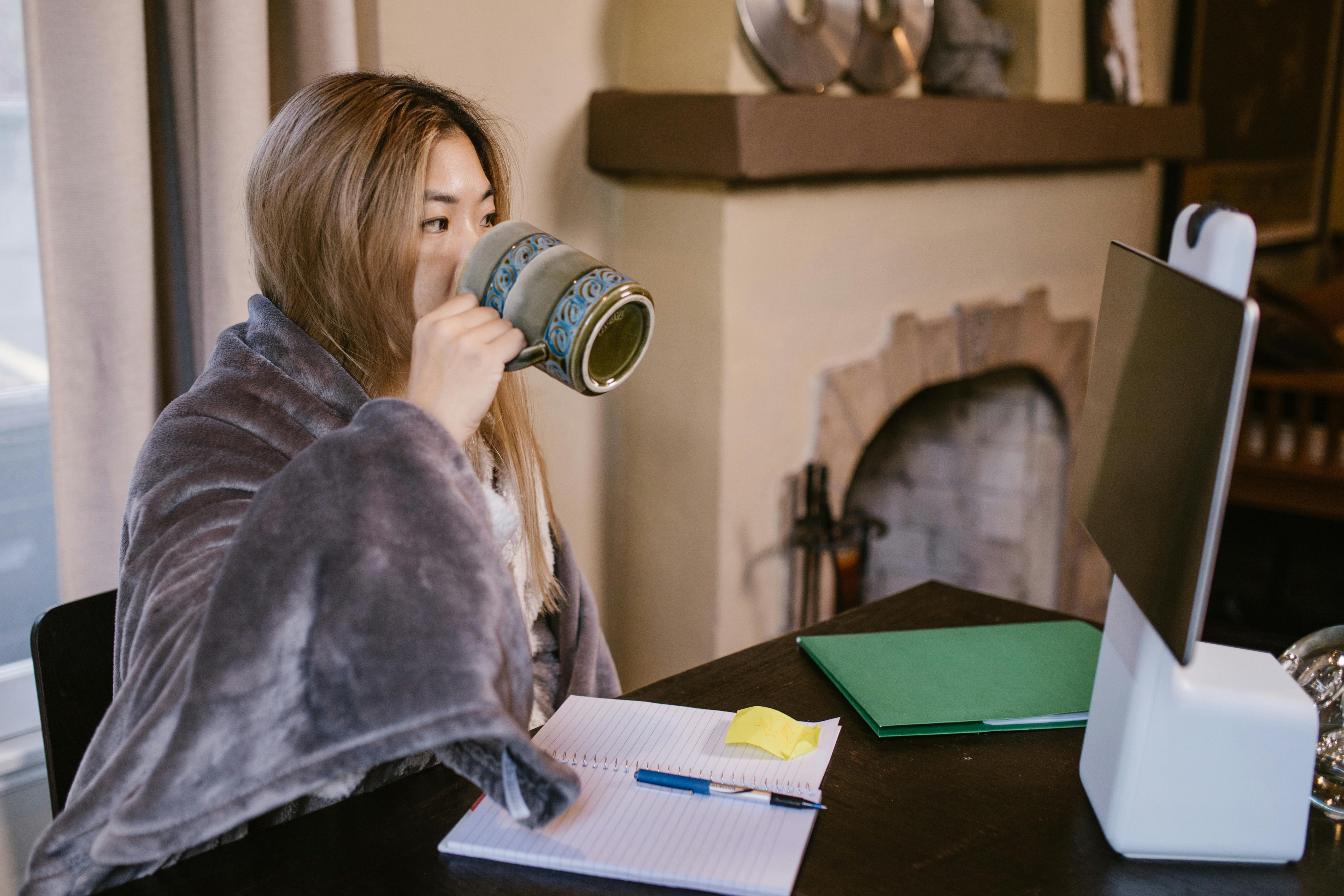A woman sitting at a desk in front of a computer, wrapped in a gray blanket, drinking from a blue patterned mug.